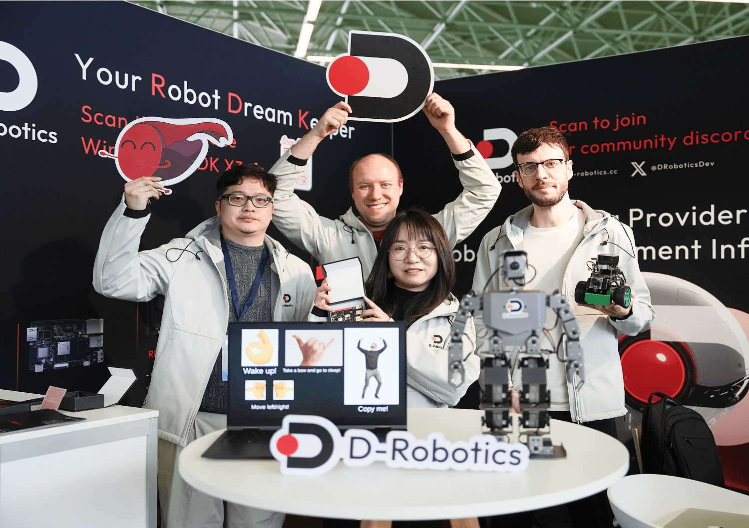 At the D-Robotics booth at DevWorld, a group of people in matching jackets hold the brand logo and small robots. There’s a humanoid robot and a computer with an interactive interface on the table, creating a lively atmosphere.