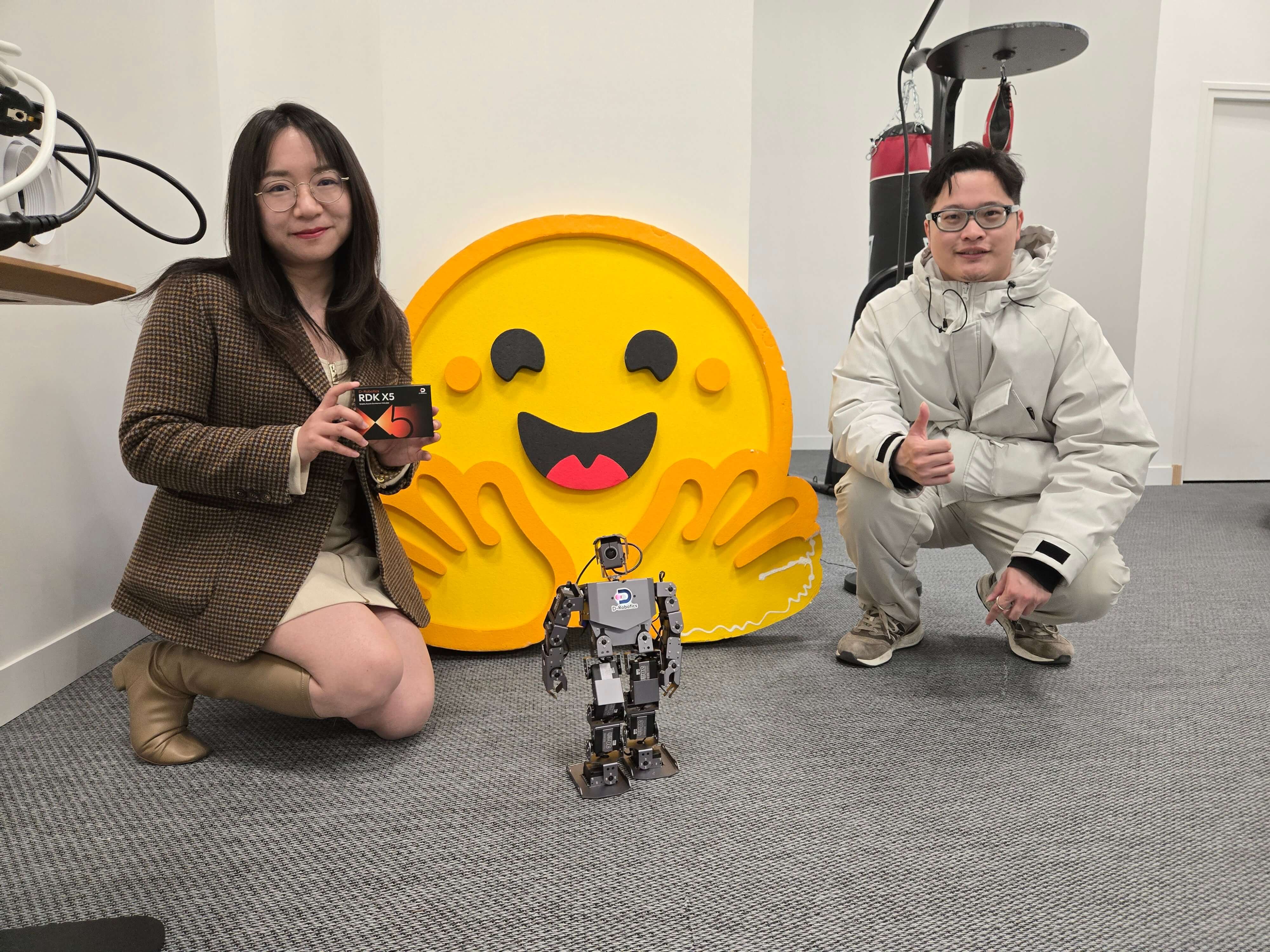 Two people pose with a small humanoid robot at Huggingface office. The woman holds “RDX X5”, with a big smiling face in the background, creating a relaxed atmosphere.