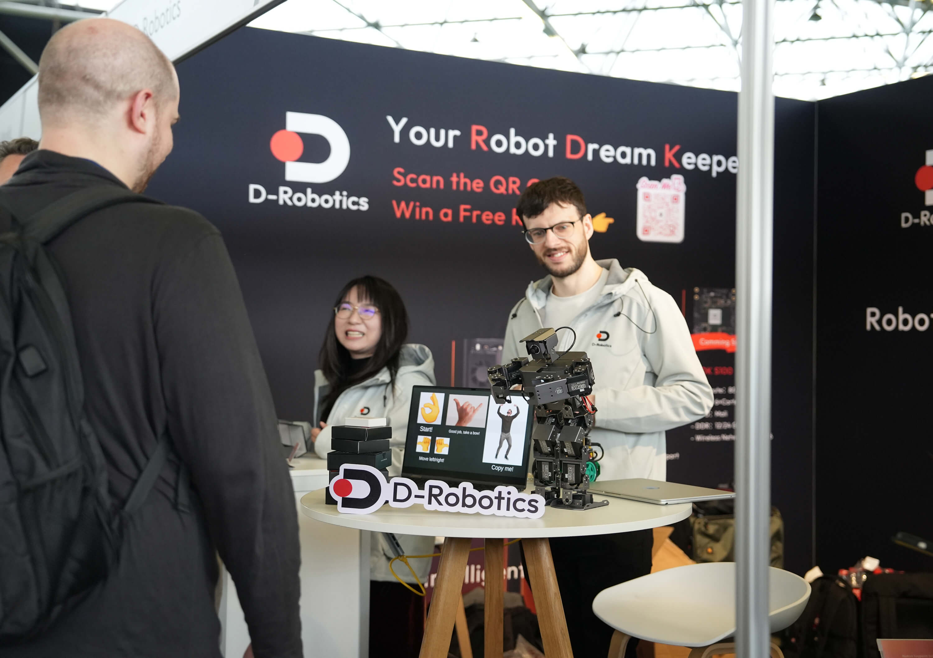 At the D-Robotics booth, two people introduce to visitors. There are humanoid robots and interactive devices on the table. With brand and event info in the background, it shows a real-time communication scene.